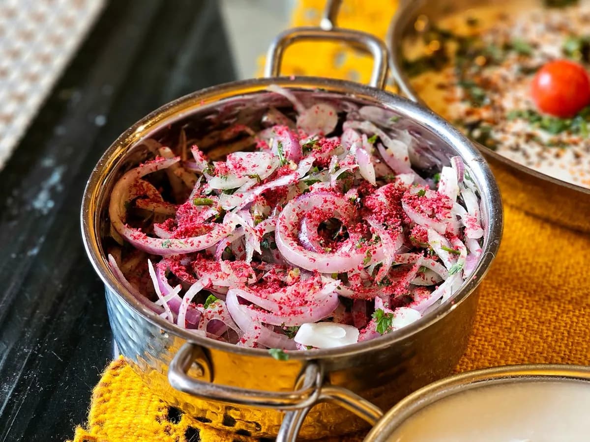 Close-up of a spiced onion salad in a brass bowl, garnished with sumac and herbs, on a vibrant table setting.
